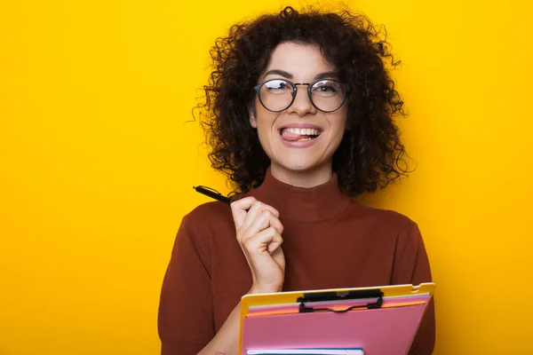 Estudiante morena de pelo rizado mirando a través de las gafas está sosteniendo un bolígrafo y algunos libros mientras posa sobre un fondo amarillo — Foto de Stock