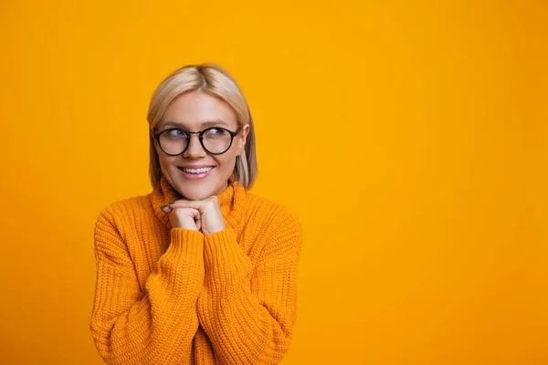 Menina loira caucasiana encantadora está olhando para o lado através de seus óculos segurando a mão perto do queixo e sorrindo alegremente no fundo amarelo com espaço livre — Fotografia de Stock