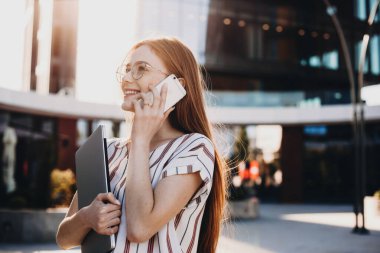 Neşeli kızıl saçlı bayan cep telefonu kullanan ve gözlüğünden bakan biriyle konuşuyor.