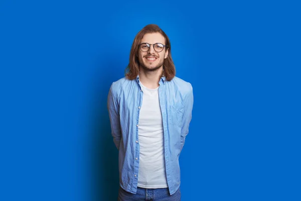 Homem caucasiano confiante com barba e cabelos longos posando em um fundo azul enquanto esconde a mão atrás das costas — Fotografia de Stock