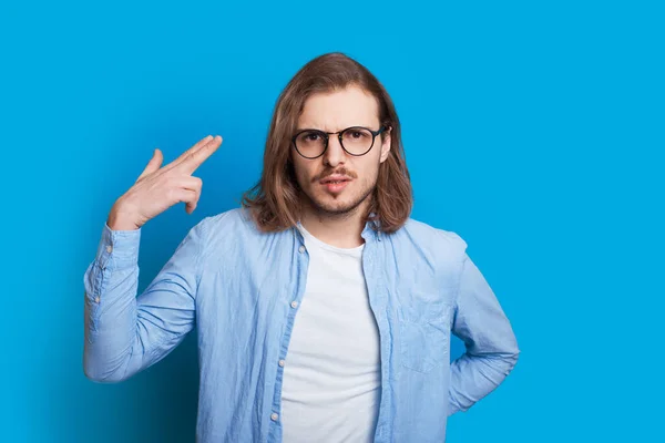 Homem caucasiano com cabelos longos e óculos gesticulando com a mão como tiro enquanto posando em um fundo azul — Fotografia de Stock