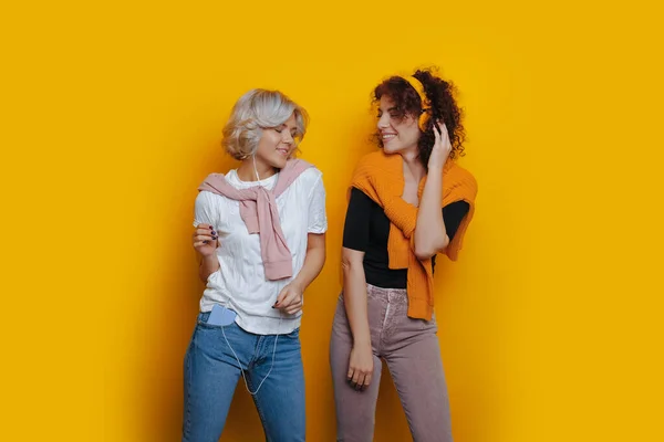 Дві радісні каскадні жінки з кучерявим волоссям слухають музику через навушники на жовтих навушниках — стокове фото