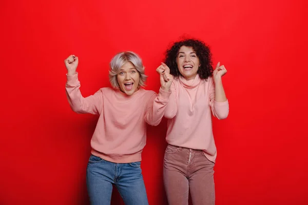 Lockige kaukasische Mädchen fühlen sich in der Nähe ihres blonden Freundes wohl, während sie auf rotem Hintergrund posieren — Stockfoto