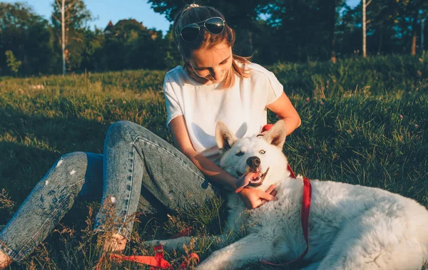 Charmante fille caucasienne avec des lunettes de soleil est d'avoir une pause assis sur l'herbe avec son chien énergétique blanc — Photo