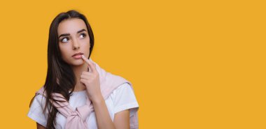 Kafkasyalı bir kız bir şey düşünürken dudaklarına dokunuyor ve boş bir arkaplanda sarı bir zeminde poz veriyor.