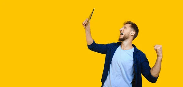 Caucásico hombre con barba y pelo rojo está mirando a su tableta y gritando ser feliz y posando sobre un fondo amarillo con espacio libre — Foto de Stock