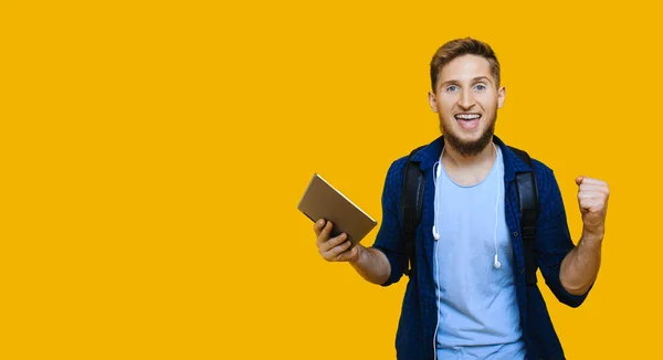 Estudiante caucásico con el pelo rojo y barba bonita está sosteniendo una tableta y usando una bolsa de la escuela, mientras que el gesto de una victoria en una pared espaciada libre amarillo — Foto de Stock