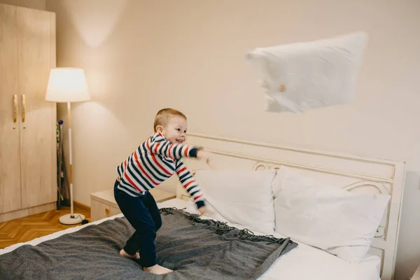 Encantador pequeño chico caucásico tirando una almohada mientras está de pie en la cama —  Fotos de Stock