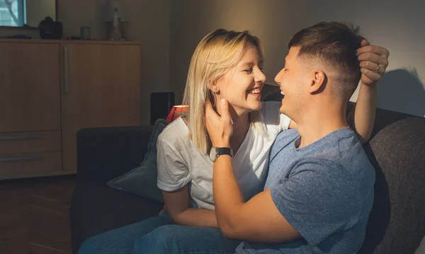 Garoto caucasiano alegre abraçando sua namorada loira enquanto está sentado no sofá e sorrindo lindamente — Fotografia de Stock