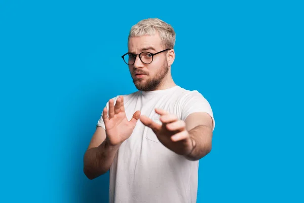 Biały mężczyzna z blond włosami i krótką brodą spogląda przez okulary i gestykuluje lękiem podczas pozowania na niebieskiej ścianie — Zdjęcie stockowe