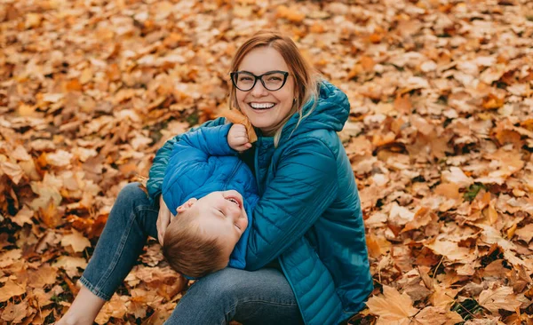 公園の紅葉の上に座っている間、赤い毛の白人の母親は息子と遊んでいます — ストック写真