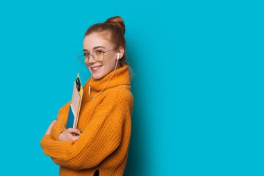 Kızıl saçlı, çilli, beyaz, çekici bir kadın elinde boş alanı olan mavi bir duvarda kitap tutarken kameraya bakıyor.