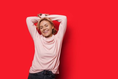 Sarı saçlı beyaz kadın ellerini başının üstüne koyup kırmızı arka planda poz veriyor.