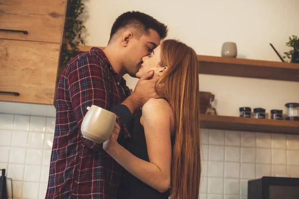 Caucasiano gengibre menina está segurando uma xícara ao ser beijada por seu amante na cozinha — Fotografia de Stock
