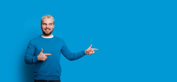 Bonito barbudo homem com cabelo loiro está apontando para a direita enquanto sorri e posando em um fundo azul com espaço em branco — Fotografia de Stock