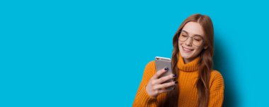 Kızıl saçlı, çilli bir kız mavi arka planda bir şeyin reklamını yaparken telefon kullanıyor.