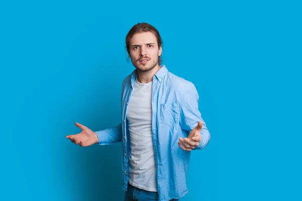 Homem caucasiano confuso com cabelos longos e barba curta está gesticulando em um fundo azul — Fotografia de Stock