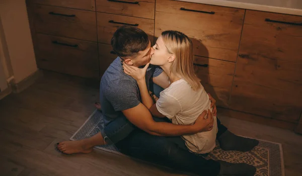 Casal caucasiano sentado no chão na cozinha e beijando uns aos outros, abraçando docemente — Fotografia de Stock