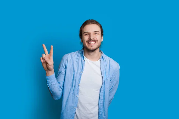 Homem caucasiano alegre com cabelos longos e barba agradável está sorrindo e gesticulando amigável em um fundo azul — Fotografia de Stock