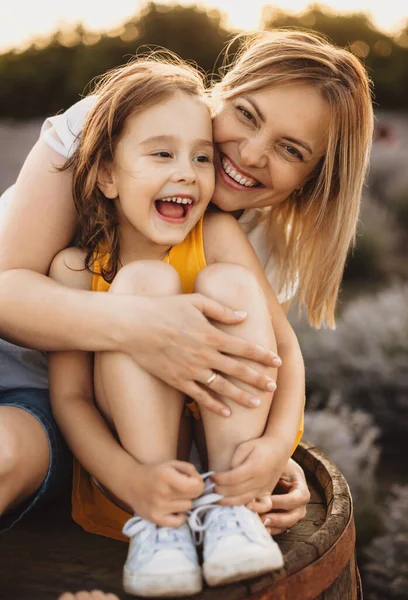 Vit flicka sitter nära sin mamma på en tunna medan hon ler och omfamnar varandra under en solig kväll — Stockfoto