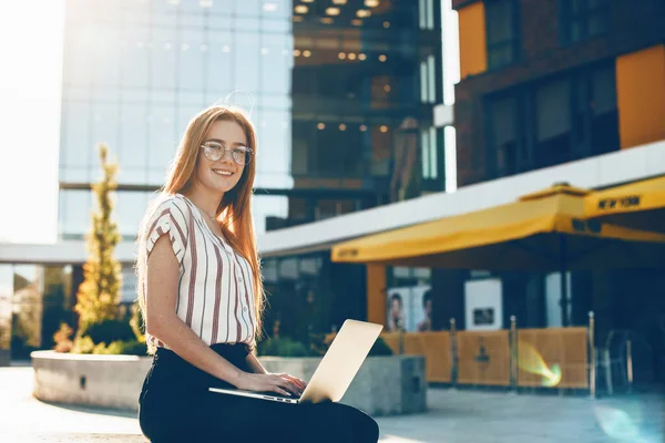 Menina de cabelos vermelhos com sardas olhando através de óculos está sentado em um banco na frente do edifício e usando um laptop — Fotografia de Stock