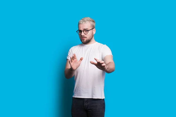 Blonde caucasian man with beard gesturing fear while looking through eyeglasses and posing on a blue background