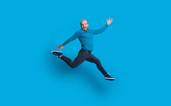 Homem caucasiano com cabelo loiro e barba vestida de azul está pulando e imitar correr em uma parede azul — Fotografia de Stock