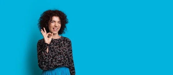 Curly cabelos caucasianos menina está gesticulando o sinal ok enquanto posando perto do espaço livre azul — Fotografia de Stock