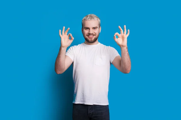 Homem caucasiano com cabelo loiro e barba bonita está gesticulando o sinal de aprovação enquanto posando em uma camiseta branca em uma parede azul — Fotografia de Stock