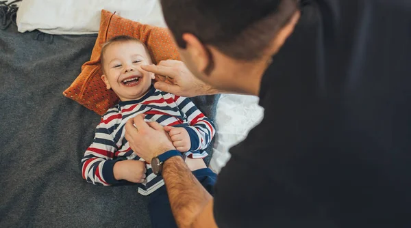 Juguetón padre caucásico hace cosquillas a su pequeño hijo que yace en el sofá y sonríe alegremente —  Fotos de Stock