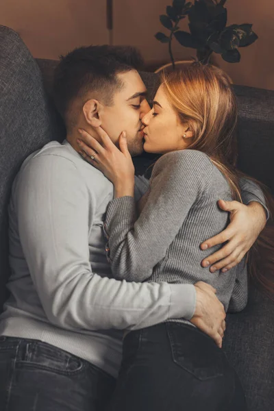 Casal caucasiano com camisolas cinza estão deitados no sofá e abraçando uns aos outros enquanto se beijam apaixonadamente — Fotografia de Stock