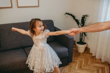 Beyaz elbiseli küçük kızıyla dans eden beyaz bir baba oturma odasında eğleniyor.