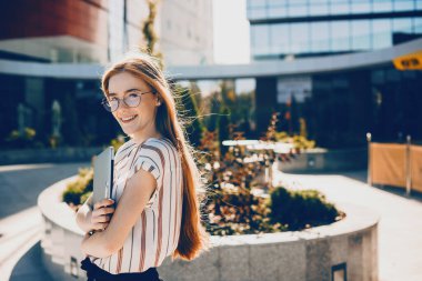 Kızıl saçlı ve çilli güzel beyaz kız bilgisayarıyla yürürken gözlüklerinden bakıyor.