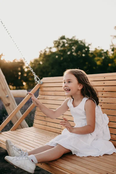 Beyaz elbiseli neşeli beyaz kız salıncakta oturuyor ve yaz akşamında eğleniyor. — Stok fotoğraf