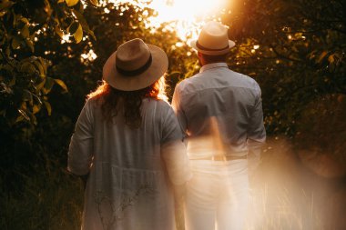 Parkta el ele yürüyen iki beyaz aşığın arka plan portresi. Birlikte gün ışığına bakıyorlar.