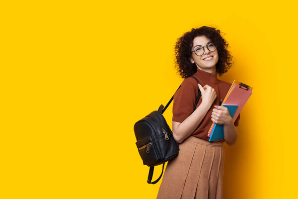 Cheerful caucasian student with a bag and some books is posing happily on a yellow background with free space