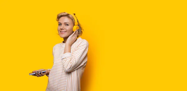 Alegre chica rubia caucásica escuchando música desde un teléfono mientras posa cerca del espacio libre amarillo — Foto de Stock