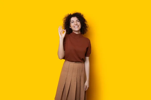 Charmerende kaukasisk pige med krøllet sort hår er gesturing på en gul baggrund, mens smilende - Stock-foto