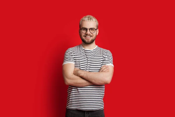 Homem caucasiano confiante com cabelo loiro e barba curta posando com as mãos cruzadas em um fundo vermelho — Fotografia de Stock