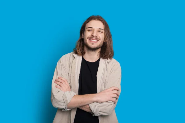 Homem caucasiano confiante com cabelos castanhos longos e barba está posando com as mãos cruzadas em uma parede azul — Fotografia de Stock