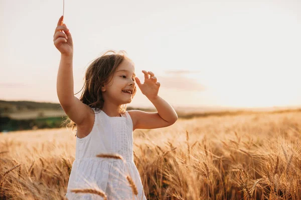 Sorrindo menina caucasiana vestida de vestido branco está andando em um campo de trigo com um sorriso alegre em saltos — Fotografia de Stock