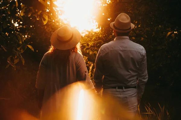 Biała para w średnim wieku w ładnych kapeluszach patrzy na zachód słońca, spacerując po ogrodzie pełnym drzew. — Zdjęcie stockowe