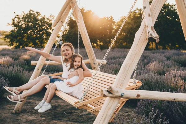 Krásná běloška matka a její malá dcera sedí v houpačce s levandulovým polem na pozadí — Stock fotografie