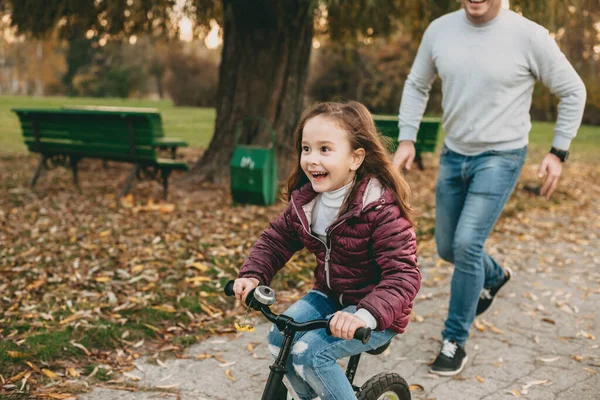 Neşeli beyaz bir baba, kızıyla parkta yürürken, kız mutlu bir şekilde bisiklete biniyor. — Stok fotoğraf