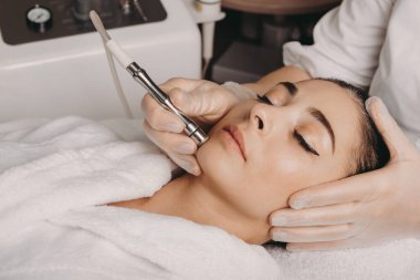 Brunette caucasian woman having a spa procedure for the skin of her face done with an apparatus by a spa worker clipart