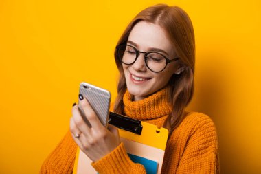 Kızıl saçlı ve çilli beyaz bir öğrenci cep telefonuna bakıyor ve elinde bazı kitaplar var.