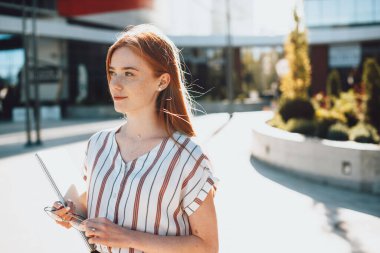 Neşeli kızıl saçlı, çilli beyaz kız dışarıda poz verirken dizüstü bilgisayarını ve gözlüklerini tutuyor.