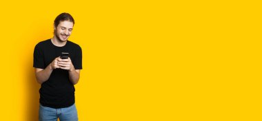 Uzun saçlı, sakallı beyaz çocuk sarı arka planda poz verirken telefonla mesajlaşıyor.