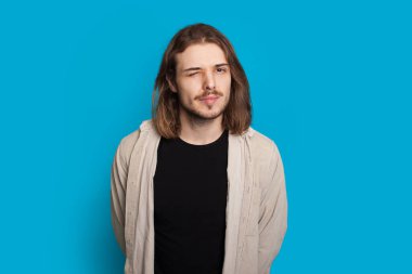 Sakallı, beyaz, uzun saçlı bir adam mavi arka planda poz verirken göz kırpıyor.