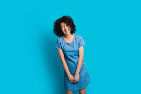 Mujer caucásica de pelo rizado posando en un bonito vestido sobre un fondo azul mientras sonríe alegremente — Foto de Stock
